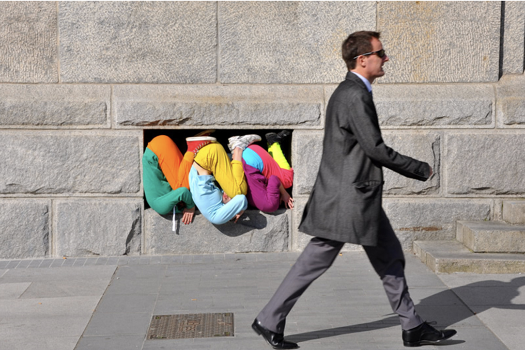 Mann geht an einer Steinwand vorbei; drei Personen in bunten Kleidern stecken in einer kleinen Öffnung.